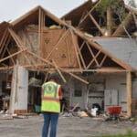 A woman standing in front of half a house with sever storm damage.