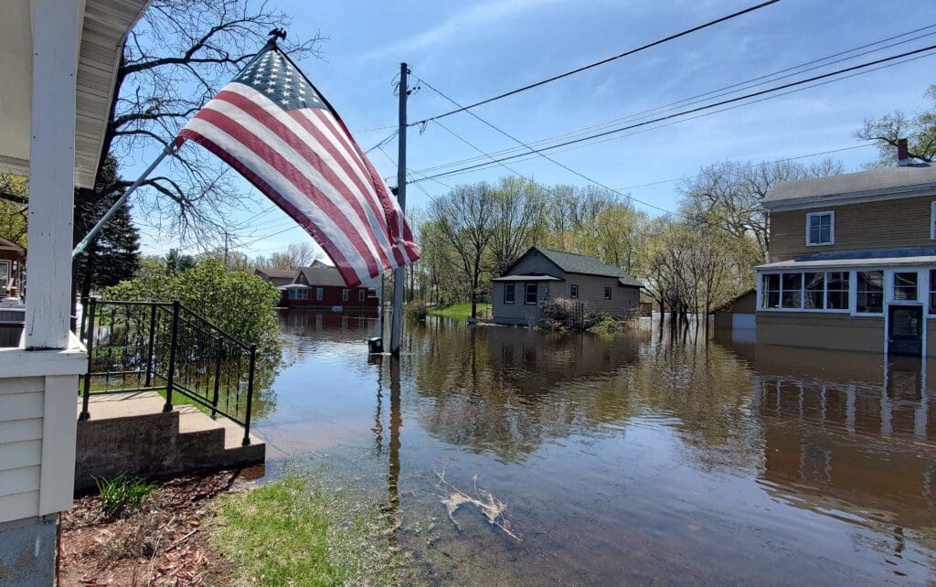 The image shows a residential area experiencing significant flooding. Several houses are partially submerged in water, reflecting a high water level that has inundated the area. In the foreground, an American flag is prominently displayed, hanging from a flagpole attached to the porch of a house. The porch itself is right at the water's edge, suggesting the floodwaters have risen quite close to the home. Power lines stretch overhead, unobstructed by the flood. The sky is clear and blue, indicating the weather is calm now, which contrasts with the disaster on the ground. Trees and greenery are visible in the background, and there is no visible movement in the water, indicating the current may be still at the moment. The scene is likely captured after a recent flood event, as the water is relatively calm and the sky is clear.